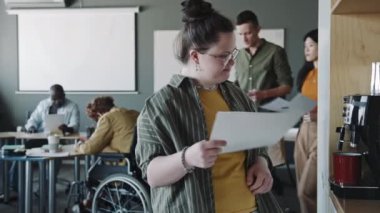 Down sendromlu genç iş kadını ofiste kahve makinesiyle fincana kahve dökerken belge okuyor.