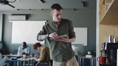Protez bacaklı genç adam dijital tablet kullanıyor, kahve makinesinden kahve alıp ofise gidiyor.