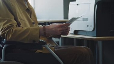 Tekerlekli sandalyedeki genç kıvırcık saçlı adam ofiste fotokopi çekerken dokümanları kontrol edip kağıda döküyor.