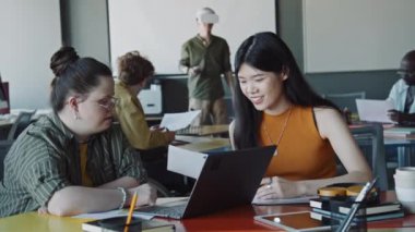 Down sendromlu genç bir iş kadını dizüstü bilgisayarda yazı yazıyor ve ofiste birlikte çalışırken Asyalı bayan meslektaşıyla konuşuyor. Arka planda VR kulaklık kullanan bir adam.