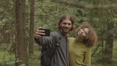Genç, neşeli bir çift telefonla selfie çekiyor ve ormanda yürüyüş yaparken fotoğraflarını kontrol ediyor.