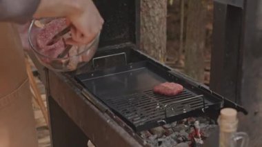 Doğadaki kamp alanında yemek pişirirken et köfteleri ve sosisleri mangalın üzerine koyan tanınmayan bir adamın ellerini yakından izleyin.