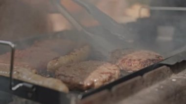 Kömür ızgarasında ızgara pişirirken köfte ve sosisleri metal maşayla çevirme görüntüsünü kapat.