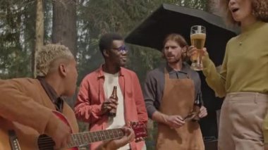 Gitar çalan ve arkadaşlarıyla birlikte şarkı söyleyen genç bir adam doğadaki barbekü partisinde eğlenirken elinde bira tutuyor.