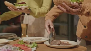 Kadın ve erkeğin açık hava barbekü partisinde masada sosisli pişirirken ekmeğe et ve sos eklerken yakın plan çekimleri.