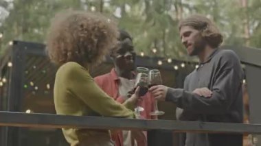 Neşeli genç arkadaşlar kırmızı şarapla kadeh kaldırıyor, sohbet ediyor ve gülüyorlar. Ormandaki kaçış evinin terasında eğleniyorlar.