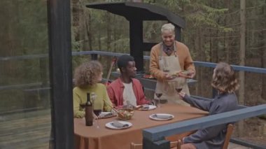 Önlüklü neşeli bir adam yanında ızgara yemek getiriyor ve arkadaşları için yemek masasına koyuyor. Ormandaki kaçış evinin terasında parti yapıyorlar.