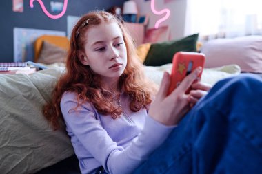 Orta sınıf, sıkılmış, kızıl saçlı bir genç kızın akıllı telefon ekranına bakarken çekilmiş bir fotoğrafı.