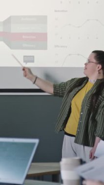 Down sendromlu genç iş kadınının dikey çekimi projeksiyon ekranındaki bilgilere ve ofis toplantısındaki ekibe sunumları gösteriyor.