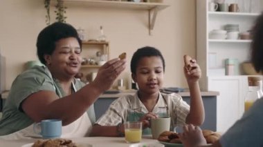 Orta yaşlı Afro-Amerikan bayanın torunlarıyla mutfak masasında oturup, taze pişmiş kurabiyelerle tezahürat yaparak, portakal suyu yiyerek ve içerek mutlu mesut zaman geçirmesi.
