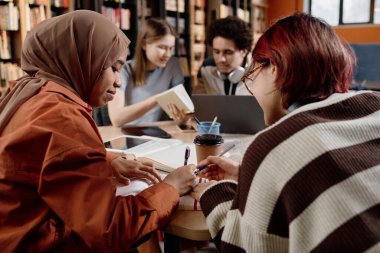 Modern okul kütüphanesinde masada oturan iki farklı kız birlikte ders çalışıp not defterine not alıyorlar.