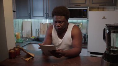 Şişman siyahi adam dijital tablette sağlıklı yemek tarifi arıyor, sonra taze meyve ve sebze getirip mutfakta yemek hazırlıyor.