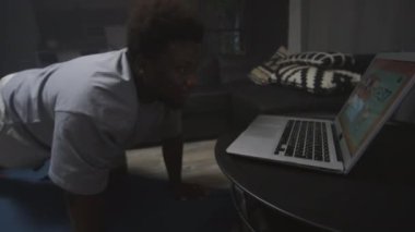 Obese siyahi adam spor giyim bölümünde dağcı egzersizi yapıyor ve evde antrenman yaparken laptopta video antrenmanı izliyor.