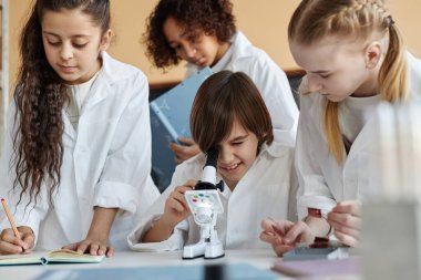 Kimya dersinde mikroskop kullanarak deney yapan laboratuvar önlüğü giyen etnik çeşitlilikte bir grup ortaokul öğrencisi.