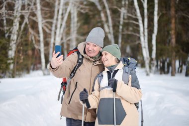 Kış günü yürüyüş yapan, akıllı telefondan selfie çeken, fotokopi çeken, orta yaşlı, beyaz bir kadın ve erkek.
