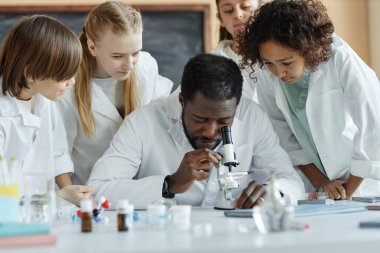 Laboratuvar önlüğü giyen genç Afrikalı bir adam ders sırasında çok ırklı erkek ve kız çocuklarına mikroskop kullanmayı öğretiyor.