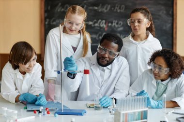 Sınıfta masada oturan genç Afrikalı Amerikalı erkek öğretmen bir grup ortaokul öğrencisine kimyasal reaksiyon gösteriyor.