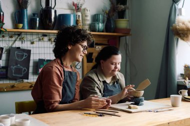 Down sendromlu genç bir kadın çömlek atölyesi sırasında zanaatkar şekillendirme kasesinin yanında oturuyor.