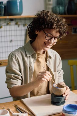 Mavi boya ve fırça kullanarak gözlük takan, el yapımı seramik kâse takan olgun, beyaz bir kadının dikey resmi.