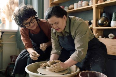 Cheerful Caucasian female artisan sitting next to woman with Down syndrome teaching her how to shape clay on potters wheel clipart