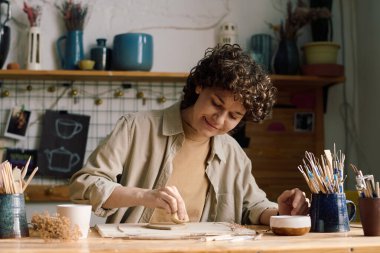 Neşeli beyaz kadın atölyede oturmuş yaprak şeklinde seramik tabaklar yapıyor.