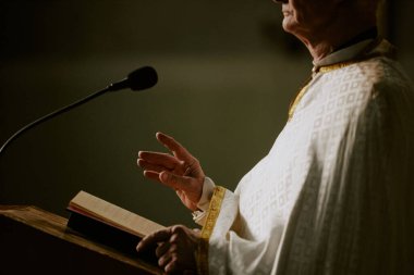 Side view crop shot of senior Caucasian Catholic priest standing at lectern with Bible book on it preaching sermon in Church clipart