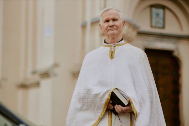 Kafkasyalı Katolik rahibin orta boy portresi elinde İncil kitabıyla dışarıda kameraya bakıyor, uzayı kopyalıyor.