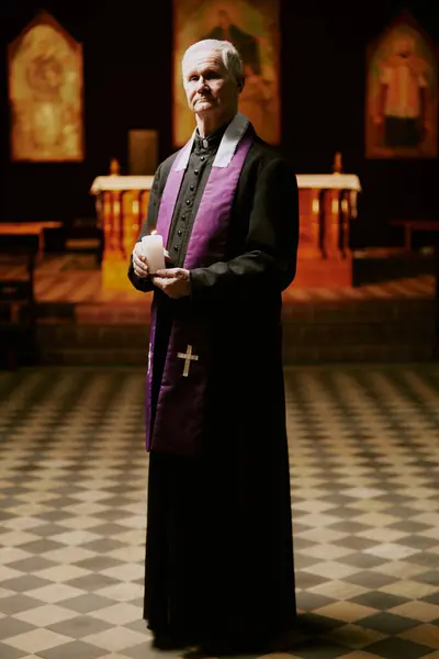 stock image Vertical long shot of senior Catholic priest wearing cassock with stole holding candle in hands standing in church looking at camera