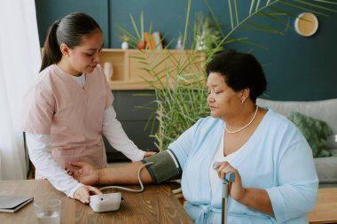 Genç hemşire, yetişkin Afrikalı Amerikalı kadınların masadaki kan basıncını ölçüyor.