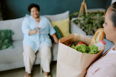 Genç hemşire, kanepede dinlenirken olgun Afrikalı Amerikalı kadın hastalara sağlıklı yiyeceklerle birlikte bakım paketi getiriyor.