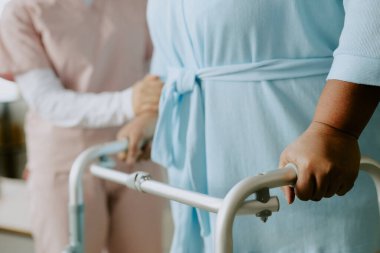 Olgun Afrikalı Amerikalı kadın hasta yürüme ve yürüme konusunda özel çaba sarf ederken hemşire kolunu tutuyor.