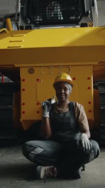 Çalışan neşeli Afrikalı Amerikalı kadın tamircinin dikey portresi. Traktör fabrikasında bağdaş kurmuş, ingiliz anahtarı tutuyor ve gülümseyerek kameraya bakıyor.
