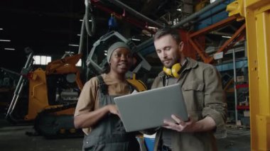 Beyaz erkek teknisyen laptopu kullanıyor ve endüstriyel fabrikada çalışan siyahi kadın işçiyle makineleri tartışıyor.