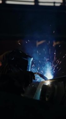 Koruyucu yüz kalkanı kaynakçısının dikey görüntüsü kaynak demiriyle gaz meşalesi ile siyah demirci atölyesinde veya endüstriyel fabrikada kıvılcımlar üretiyor.