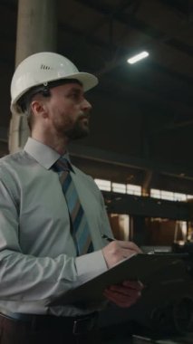 Formalite ve güvenlik başlığı takmış fabrika müfettişinin dikey çekimi. Üretim sürecini izliyor ve panoya notlar yazıyor.