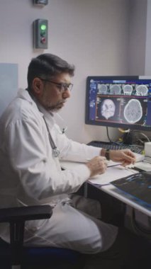 Orta yaşlı erkek nörolojistin MRI kontrol odasında oturup filmdeki beyin taramasını kaldırırken, tomografi görüntülerini analiz ederken ve yorumlarken orta boy dikey yavaş çekimi.