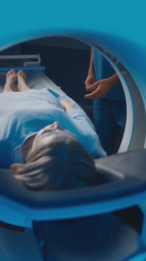 MRI kapsülünün içinde hastane önlüklü bir kadının tarama yatağında klinikte teşhisi için hazır yattığı dikey görüntü. Erkek hemşire hastaların elini tutuyor ve destek işareti gösteriyor.