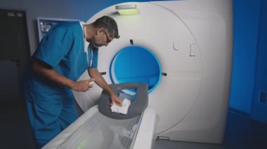 Yetişkin erkek çok ırklı görüntüleme uzmanı antiseptik spreyle dezenfekte edip kağıt peçete kullanarak MRI cihazlarını hazırlıyor.