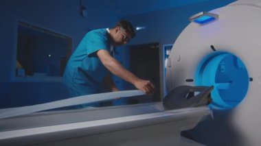 Yetişkin erkek görüntüleme uzmanının hastaların masasına yeni bir tek kullanımlık sağlık tablosu koyarak MRI cihazlarını prosedür için hazırlarken orta yavaş çekimi.