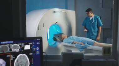 Kafkasyalı bir kadının tarama yatağında yatıp PET işlemine hazır bir şekilde görüntüsü. Erkek çok ırklı radyoloji uzmanı kadın hastayı uzaktan kumandayla MRI makinesine sokuyor.