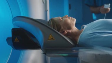 Hasta masasında yatan orta yaşlı beyaz kadının yavaş yavaş yakın plan görüntüleri uzaktan kumandayla kontrol edilen radyoloji uzmanı tarafından kontrol edilen MRI makinesine taşınıyor.
