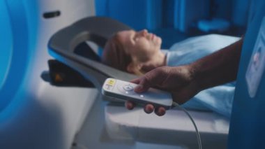Hasta izleme sistemini kullanarak uzaktan kumandalı MRI cihazı kullanan isimsiz bir erkek radyologun yavaş yavaş el çekimi. Tarayıcı yatağında yatan kadın teşhis makinesine giriyor.