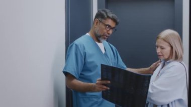 Çok ırklı erkek pratisyen hekimin klinikte bekleyen orta yaşlı beyaz kadın hastaya film taraması göstermesi MRI sonuçlarını duyurması ve şok olmuş kadını desteklemesi.