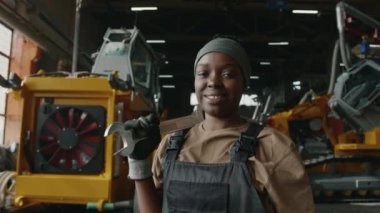 İngiliz anahtarı tutan ve makine fabrikasında gülümseyerek kameraya poz veren neşeli siyah kadın tamirci portresi