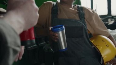 İş kıyafetleri içindeki kadın teknisyenin, kahve molası sırasında iş arkadaşıyla konuşurken elinde baret ve termos bardakla orta bölüm yakın çekimi.