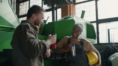 İş kıyafetlerindeki genç Afrikalı Amerikalı teknisyen termostan kahve içiyor ve endüstriyel fabrikada tatilde beyaz erkek iş arkadaşıyla sohbet ediyor.