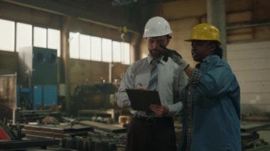 Beyaz erkek fabrika mühendisi. Formalite ve başlık takıyor. Pano tutuyor ve siyah kadın teknisyenlere iş görevlerini anlatıyor.