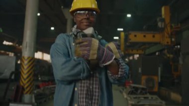 Koruyucu iş kıyafetlerinde çalışan, kollarını kavuşturup endüstriyel fabrikada poz verirken kameraya gülümseyen kendinden emin kadın teknisyenin zoom portresi.