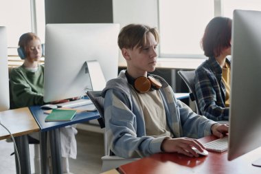 Modern sınıfın masalarında oturan üç ergen ders sırasında modern bilgisayarları kullanıyorlar.