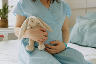 Young pregnant woman sitting on hospital bed while she holding plush bunny and hugging her belly clipart